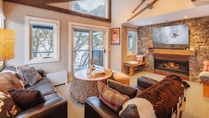 A cozy living room with a stone fireplace, leather sofas, a round wooden coffee table, and a large TV mounted on the wall. - The Stables Resort Perisher - Open All Year (Perisher Valley)