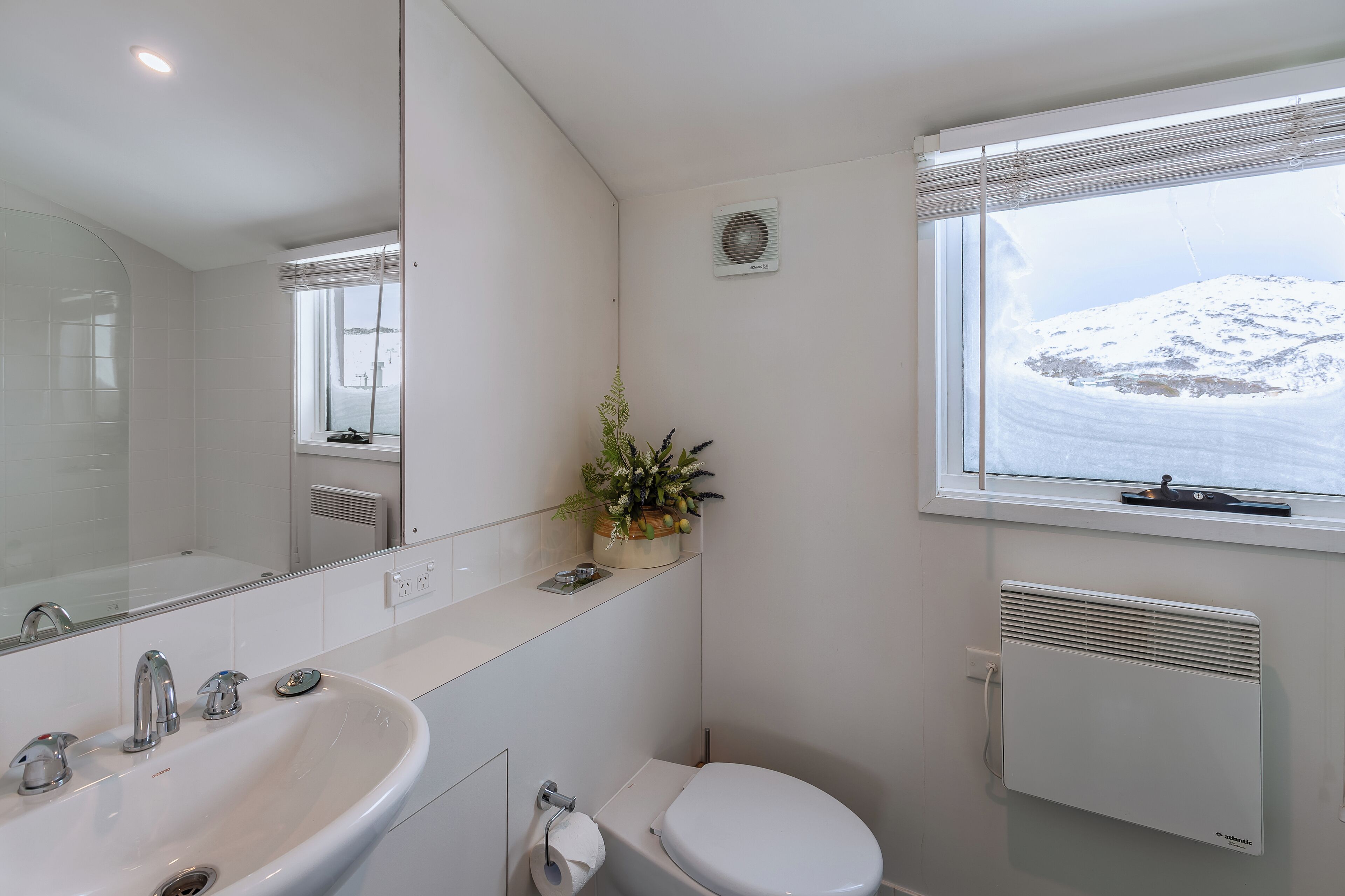 A bathroom with a white sink, a toilet, a mirror, and a window with a view of a snowy landscape.