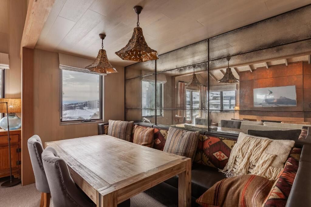 Una sala de estar acogedora con una mesa de comedor de madera, asientos con cojines y grandes ventanales con vistas al exterior.