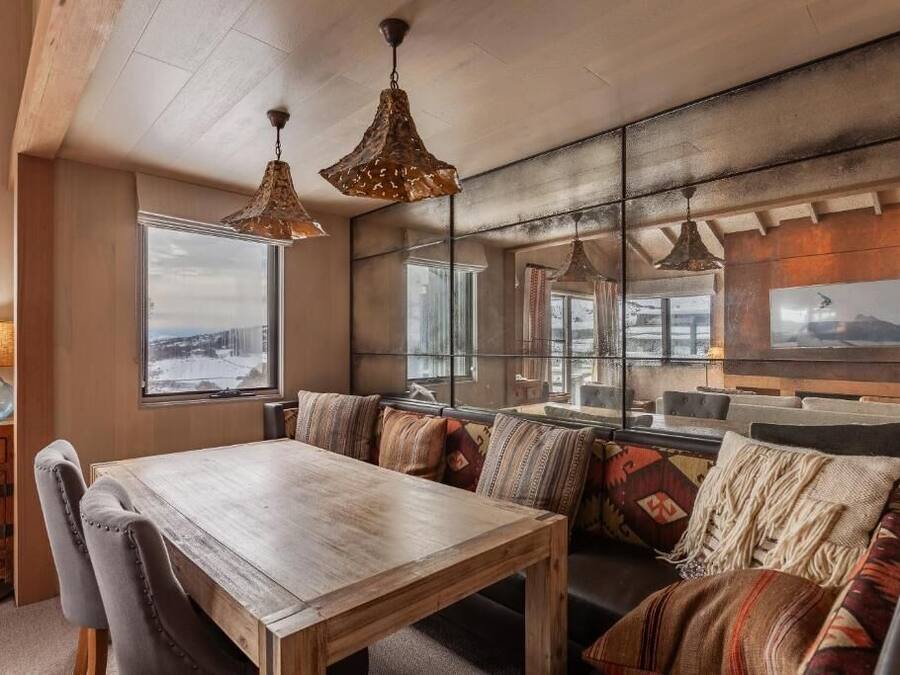 Una sala de estar acogedora con una mesa de comedor de madera, asientos con cojines y grandes ventanales con vistas al exterior.