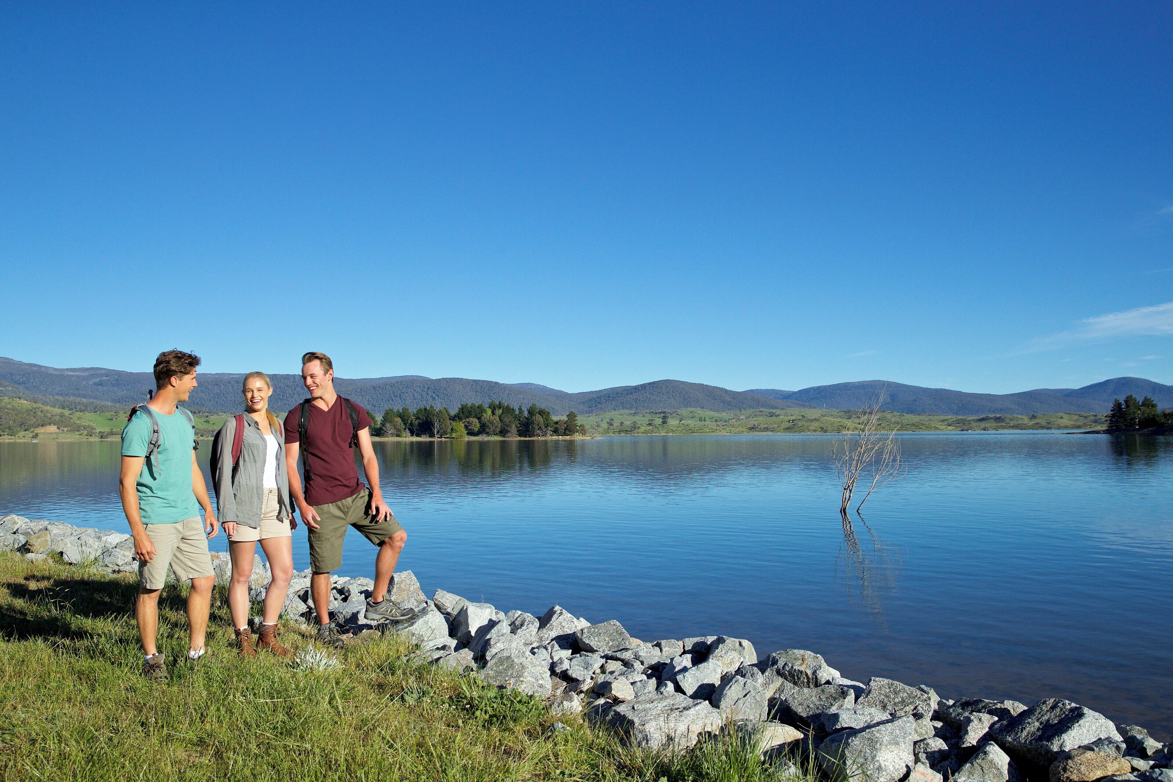 Drei Personen stehen an einem See mit felsigem Ufer und Bergen im Hintergrund.