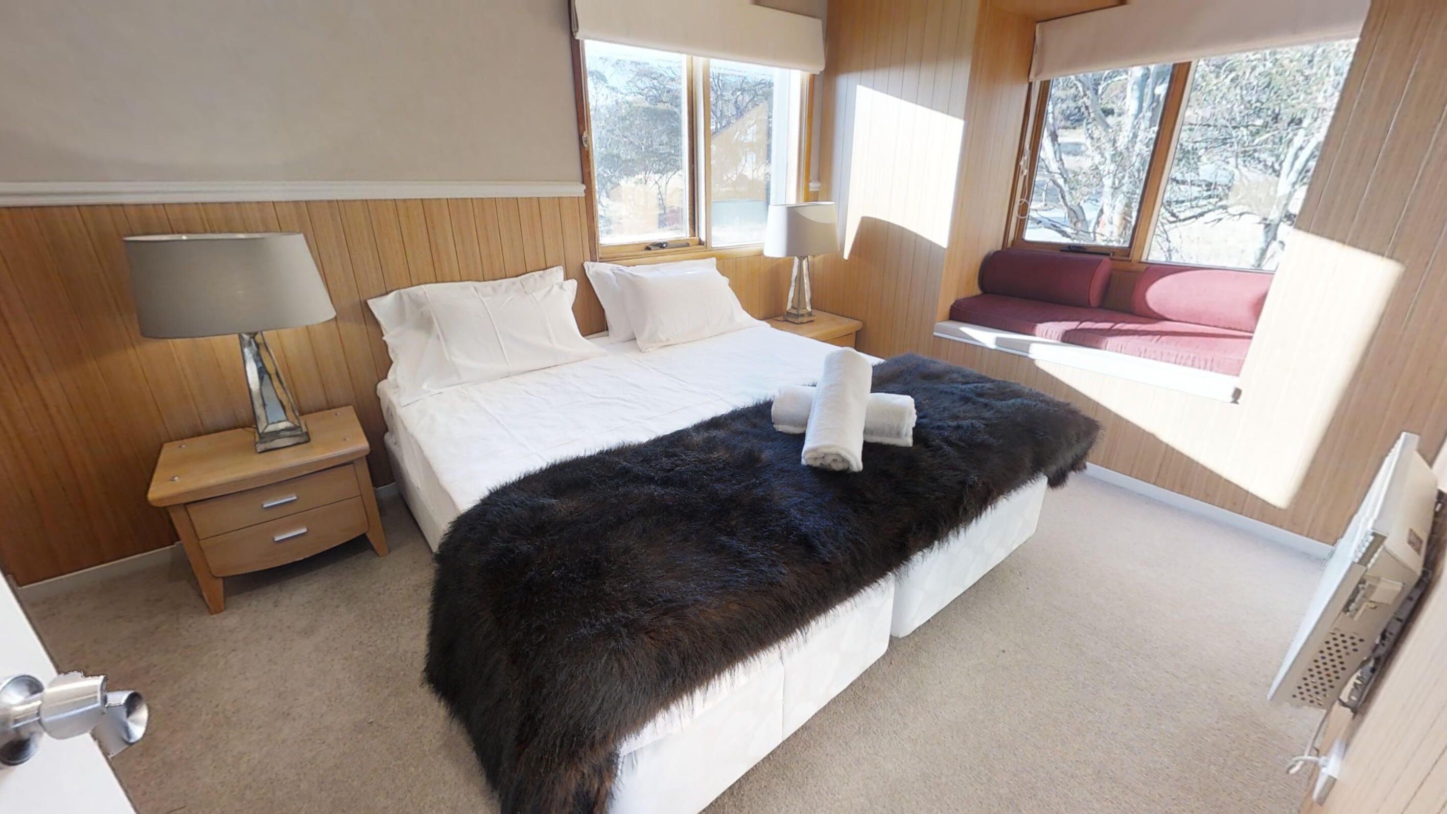 A bedroom with a large bed, a nightstand, a lamp, a window with a view of trees, and a sofa in the corner.
