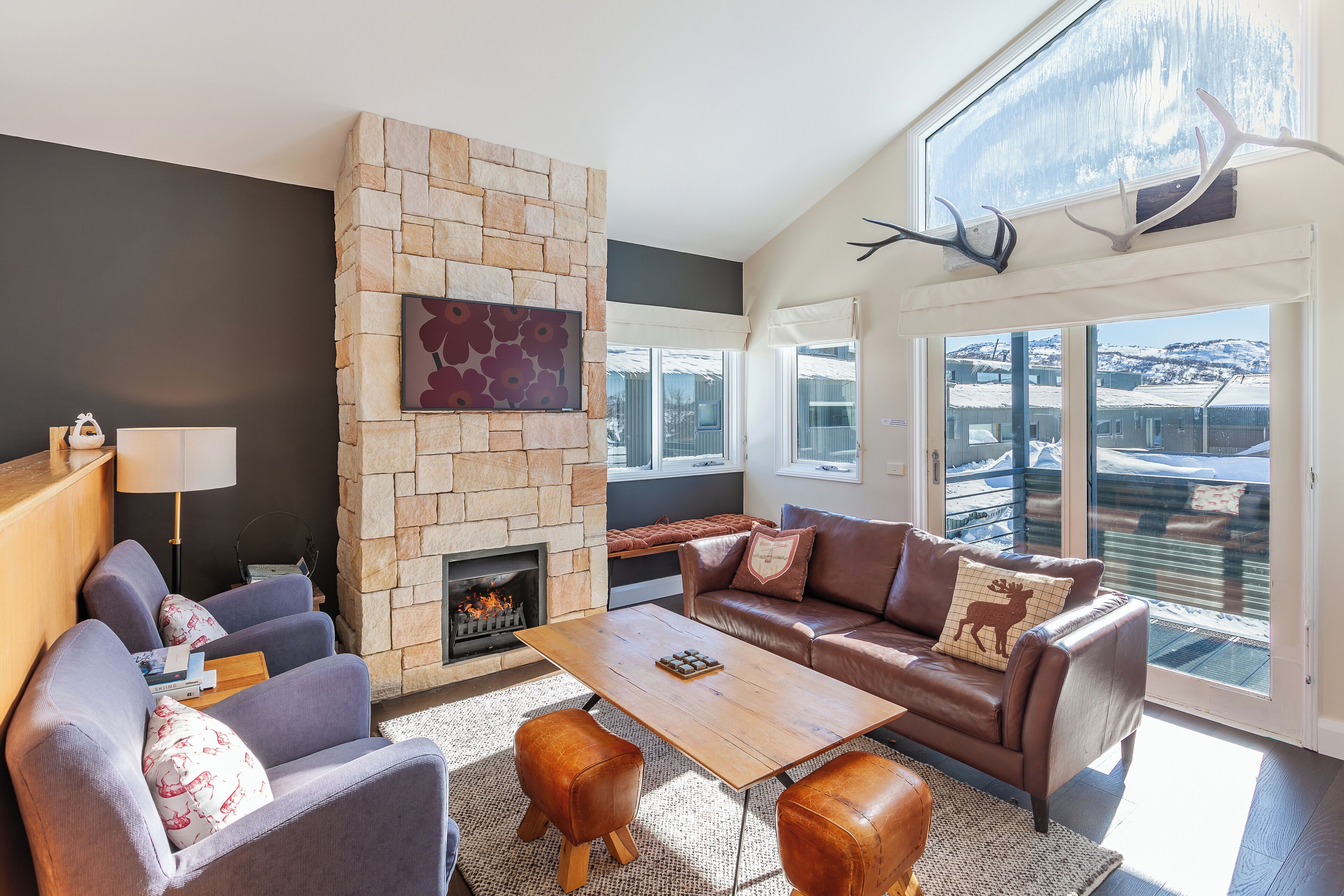 A modern living room with a stone fireplace, leather sofas, a wooden coffee table, and a large window with a view of a snowy landscape.