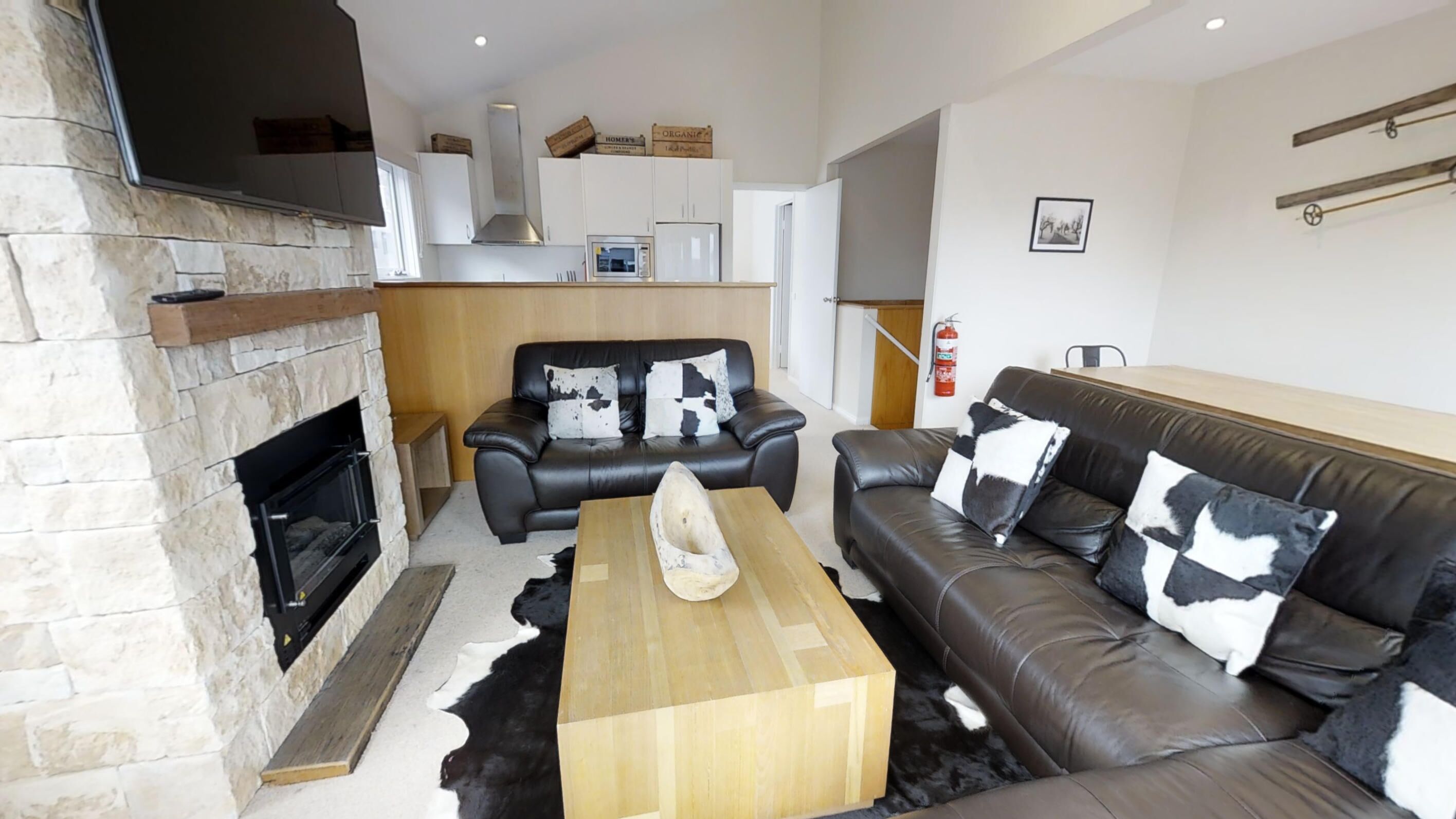 A modern living room with a stone fireplace, leather sofas, a wooden coffee table, and a kitchen in the background.
