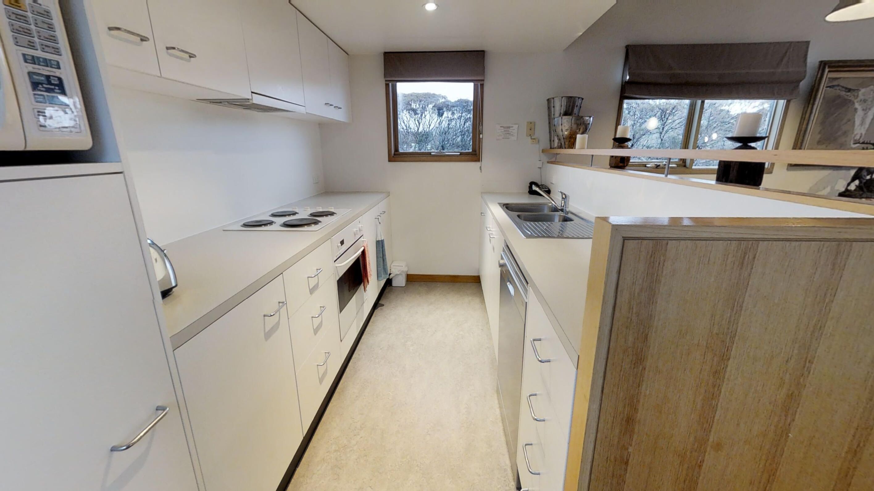 Une cuisine de taille réduite, avec des meubles blancs, une cuisinière, un évier et une fenêtre donnant sur les arbres.