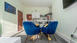 A modern kitchen with white cabinets, a built-in stove, and a microwave. There are two blue chairs with gold legs and a white table in the foreground. - 1 Bed Flat Wanstead E11 2PF | Save 10% LongStay | Contractors | Wi-Fi | Sleeps4 | Parking (Greater London)