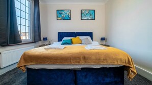 A hotel room with a large bed, a window with curtains, two framed artworks on the wall, and a bedside table with a lamp. - 1 Bed Flat Wanstead E11 2PF | Save 10% LongStay | Contractors | Wi-Fi | Sleeps4 | Parking (Greater London)