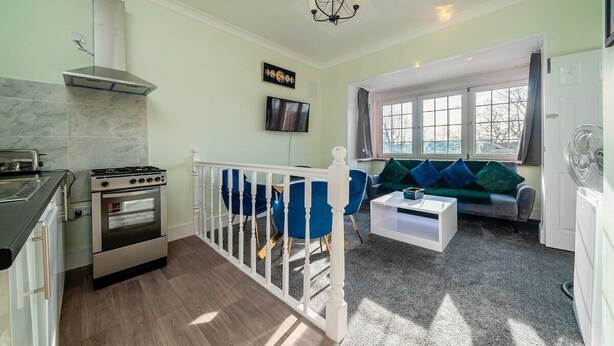A modern kitchen with a stove, oven, and a chimney. There is a dining area with blue chairs and a white coffee table. A TV is mounted on the wall, and a window provides natural light. - 1 Bed Flat Wanstead E11 2PF | Save 10% LongStay | Contractors | Wi-Fi | Sleeps4 | Parking (Greater London)