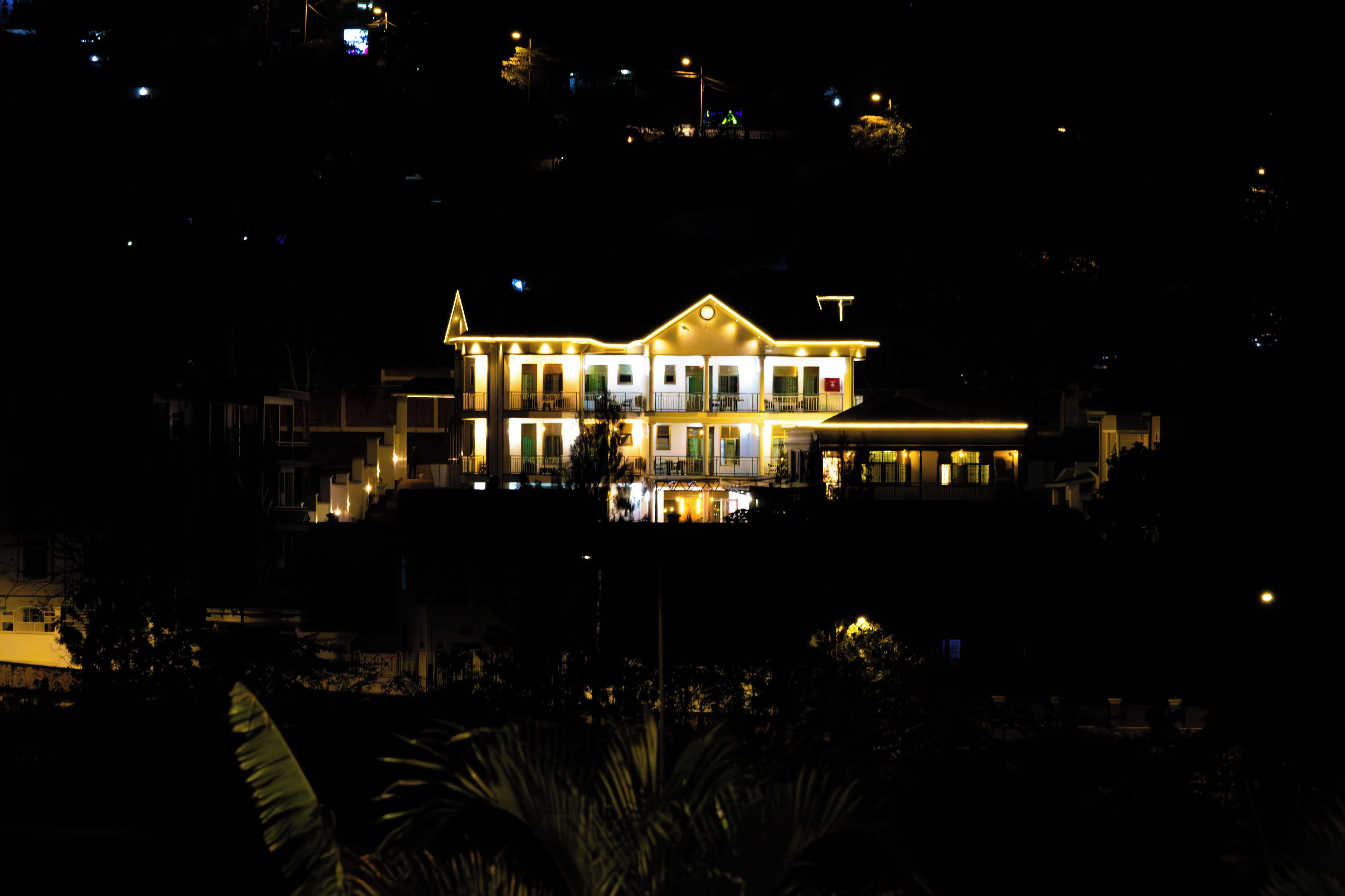 A large, illuminated building with multiple levels and balconies, surrounded by dark foliage.