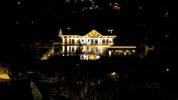 A large, illuminated building with multiple levels and balconies, surrounded by dark foliage.