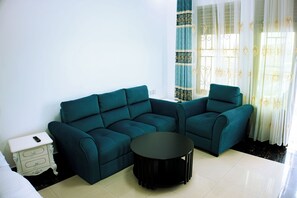 A teal sectional sofa, a small round table, and a white bedside table with a remote control. - ROYAL CROWN HOTEL  (Kigali)