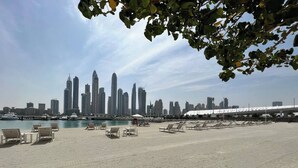 A beach with lounge chairs and a marina in the background. - Marina Vista Residences - Dubai Marina (Dubai)