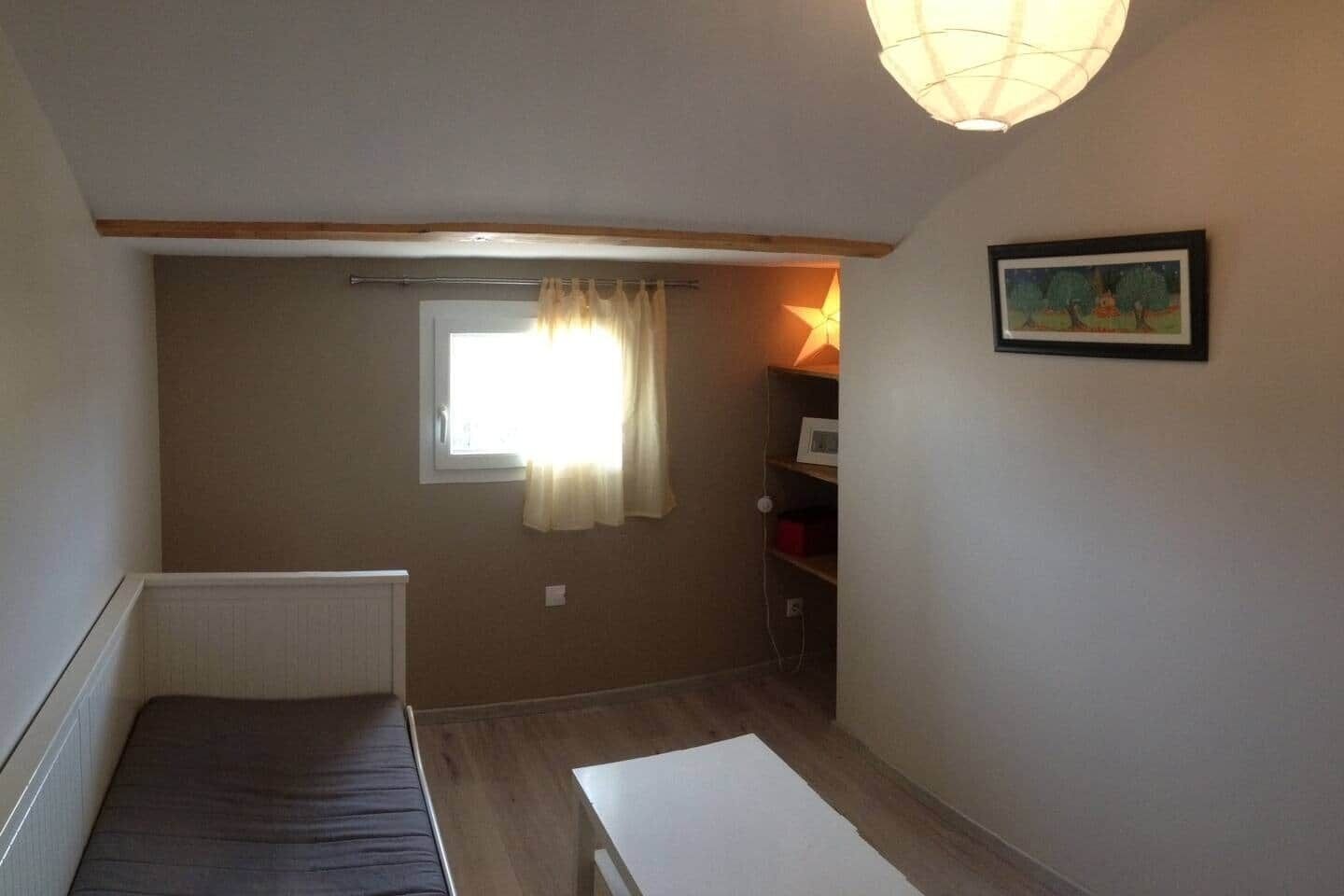 Een kleine, eenvoudig ingerichte kamer met een raam, een radiator, een plank met spullen en een fotokader aan de muur.