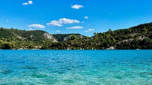 Vista despejada de un mar turquesa con patrones submarinos visibles, un paisaje montañoso con vegetación densa y un cielo despejado.