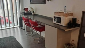 Ein modernes Hotelzimmer mit einer Küchenzeile, einer kleinen Essecke mit roten Stühlen, einem großen Fenster mit Jalousien und einem Willkommensschild an der Wand.
