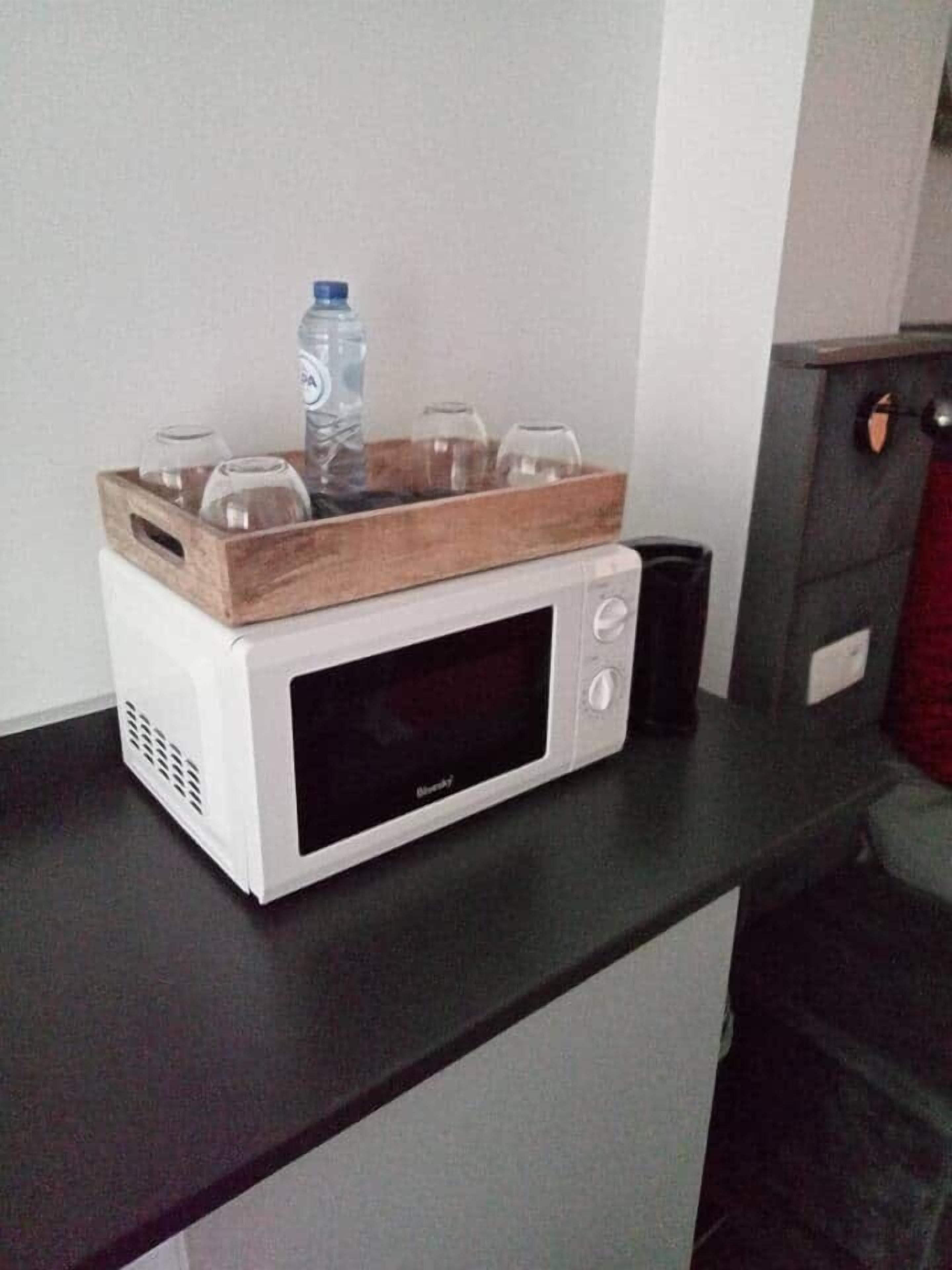 Un forno a microonde bianco su un piano di lavoro nero con un vassoio di legno sopra, contenente una bottiglia d'acqua e dei bicchieri.