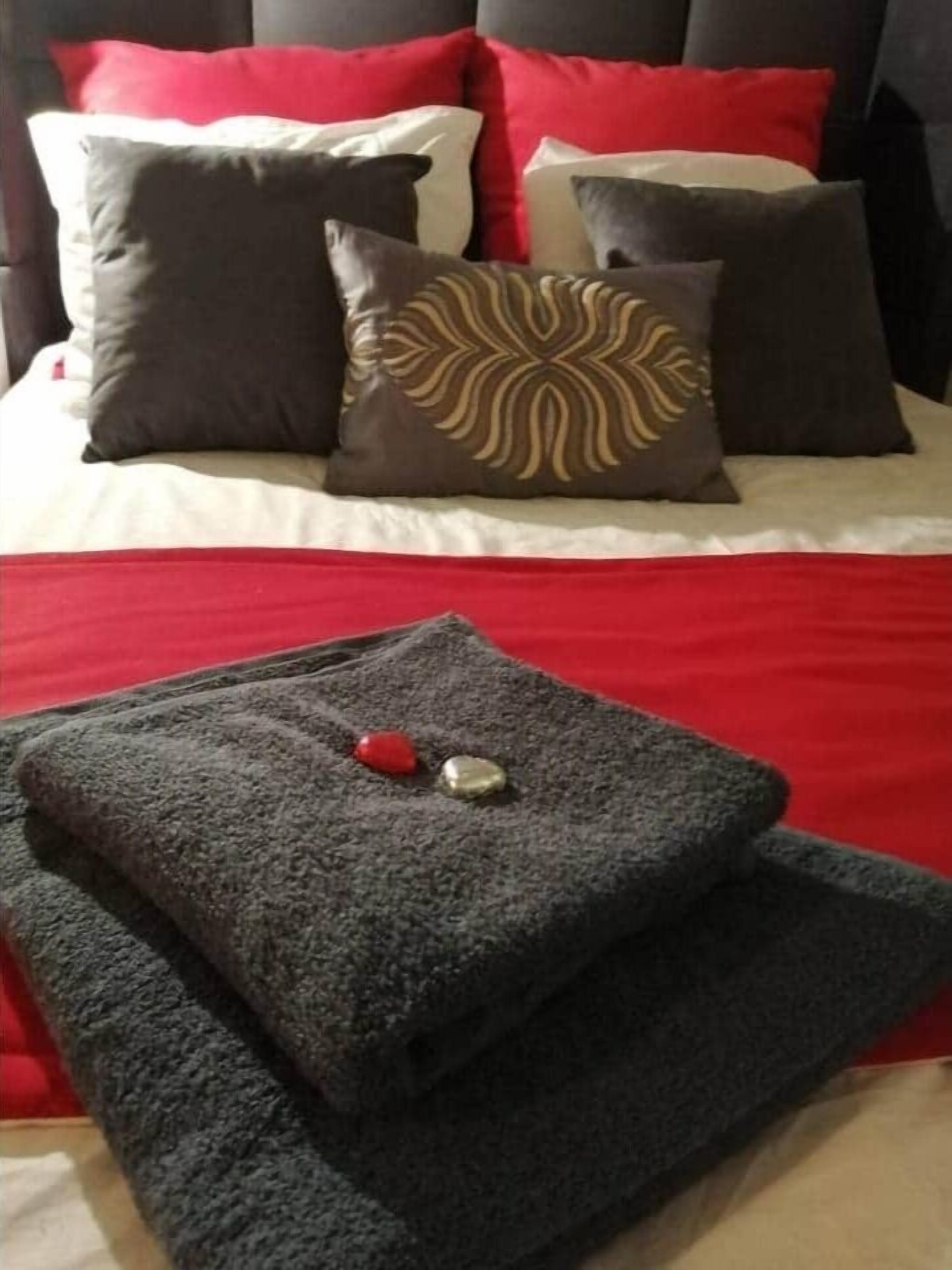 Una cama con una colcha roja y blanca, varios cojines de distintos colores y estampados, y una toalla gris oscuro doblada con un objeto rojo y dorado encima.