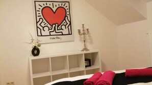 Ein Schlafzimmer mit einem Bett, einem Nachttisch mit einem Kerzenleuchter, einem gerahmten Kunstwerk darüber und einer Vase mit Blumen.