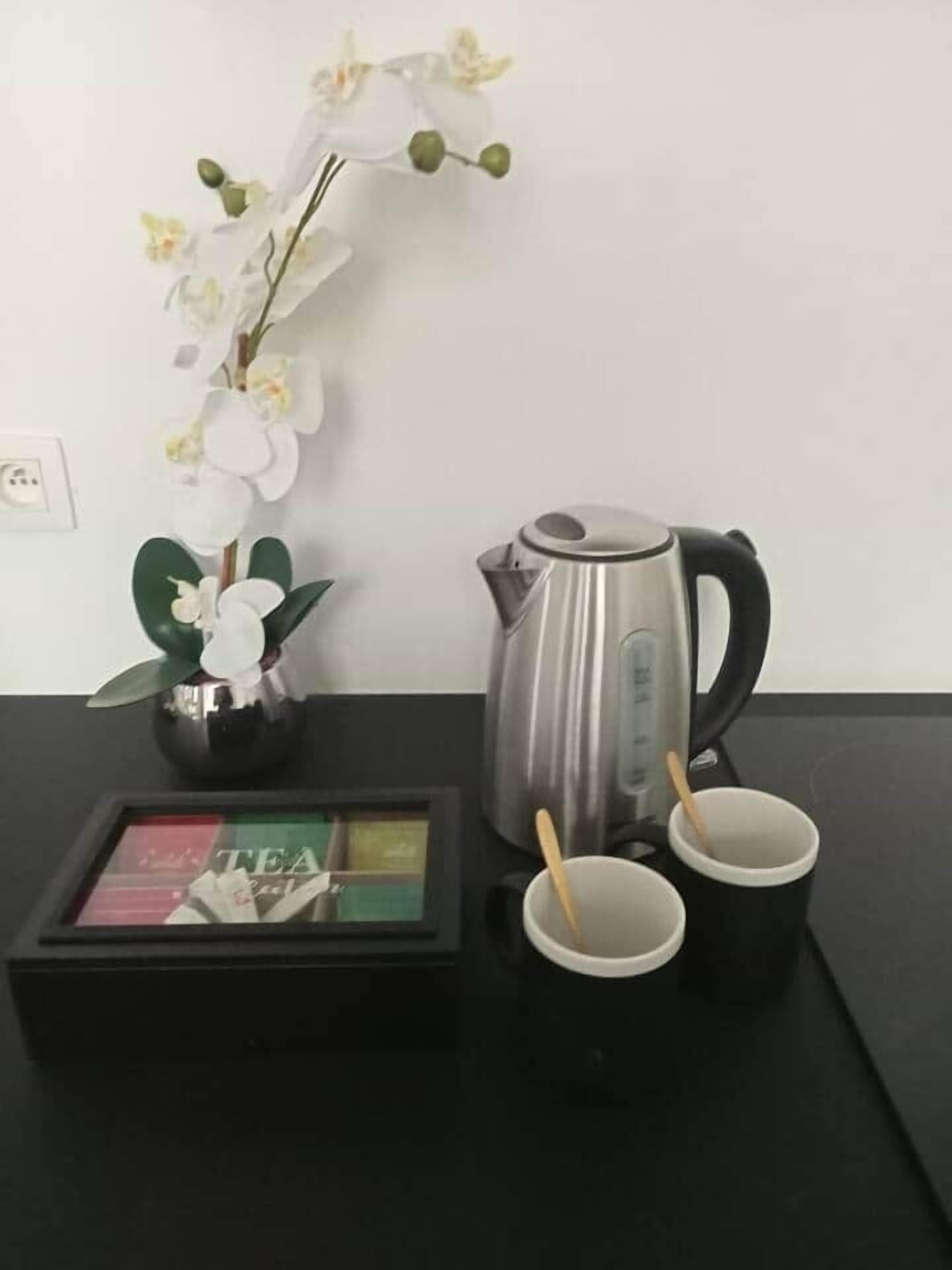 Una mesa negra con un hervidor eléctrico plateado, dos tazas negras con cucharas de madera, un expositor de té enmarcado y un florero con orquídeas blancas.
