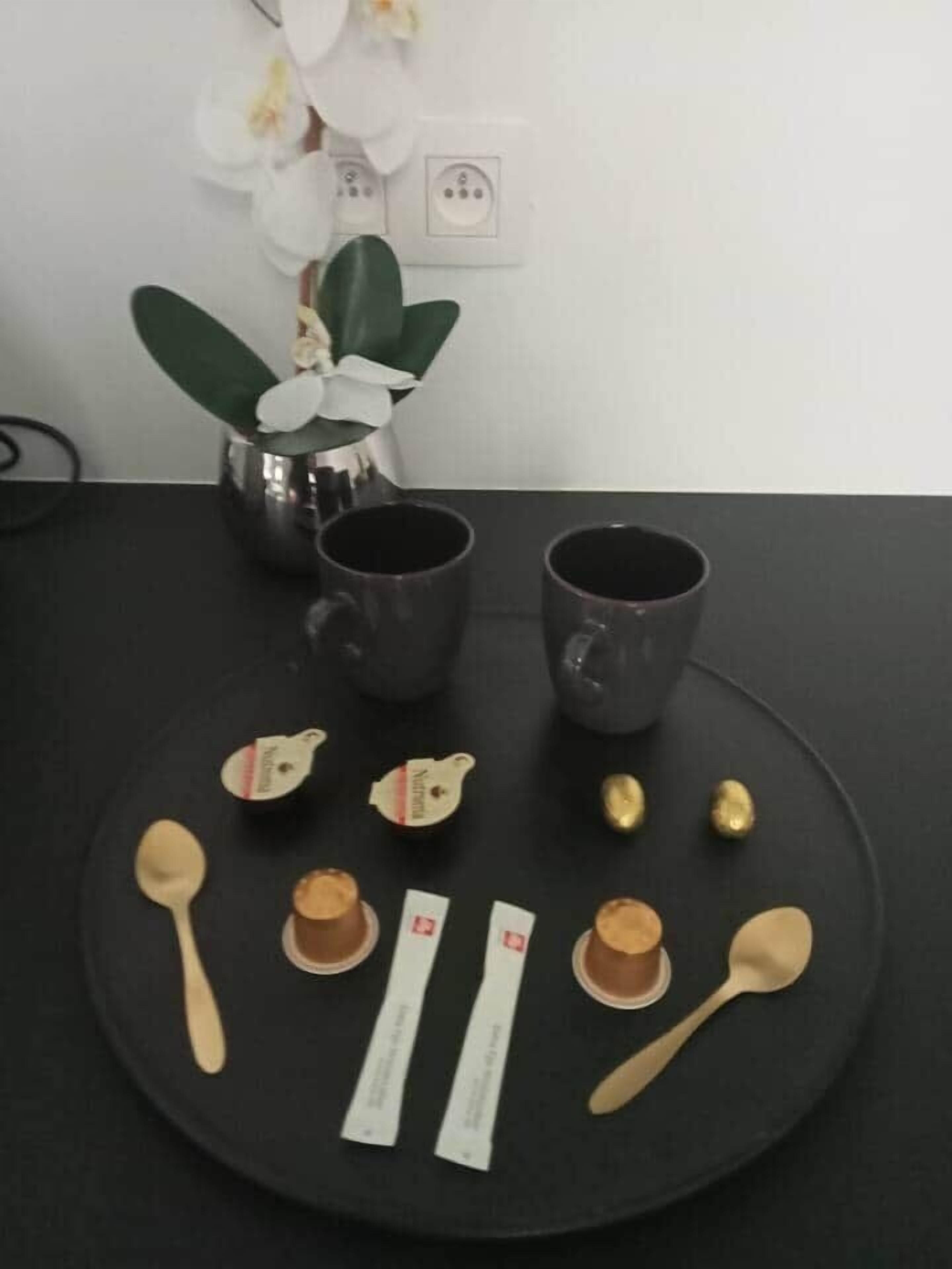 Une table avec deux tasses foncées, une cuillère, deux sachets et une petite plante aux fleurs blanches.