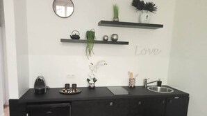 Un comptoir de cuisine noir avec une bouilloire, des tasses et un évier. Des étagères se trouvent au-dessus, ornées d’objets décoratifs, et un miroir est accroché au mur.