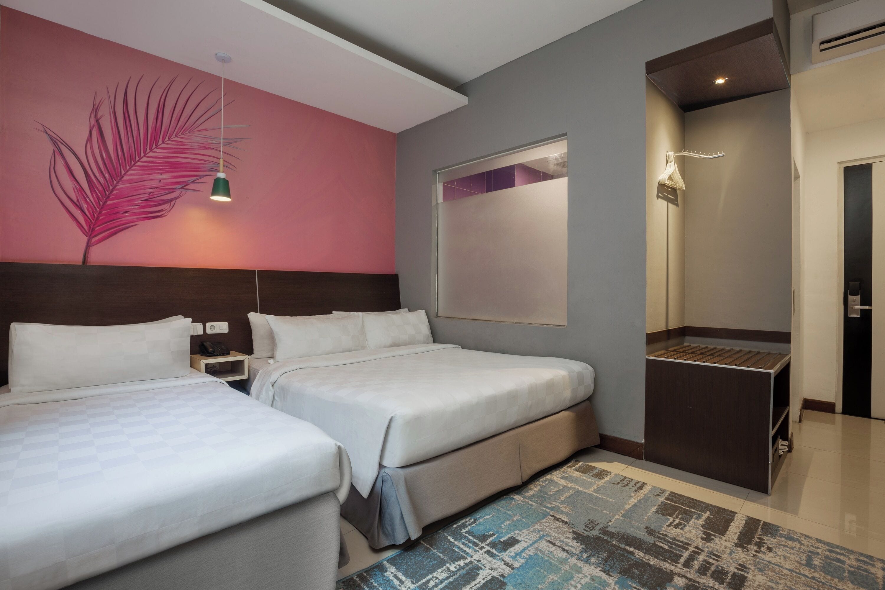 A hotel room with two beds, a large mirror, a nightstand, a wall-mounted lamp, and a small wooden shelf.