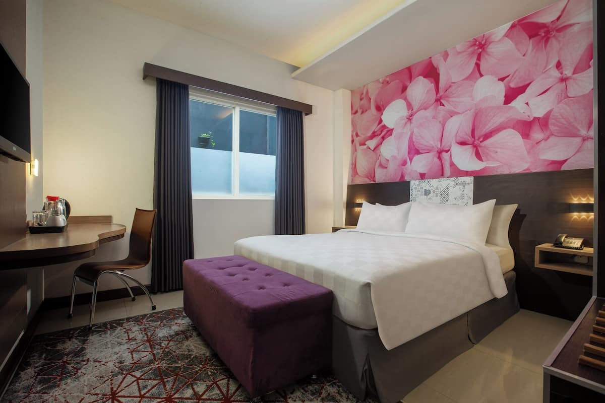 Ein Hotelzimmer mit Bett, Schreibtisch, Sessel, violettem Hocker, großem Blumenwandbild und einem Fenster mit Vorhängen.