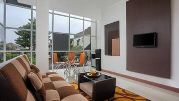 Una sala de estar moderna con un sofá, una mesa de centro con fruta, un televisor montado en la pared y ventanas grandes con vistas al exterior.