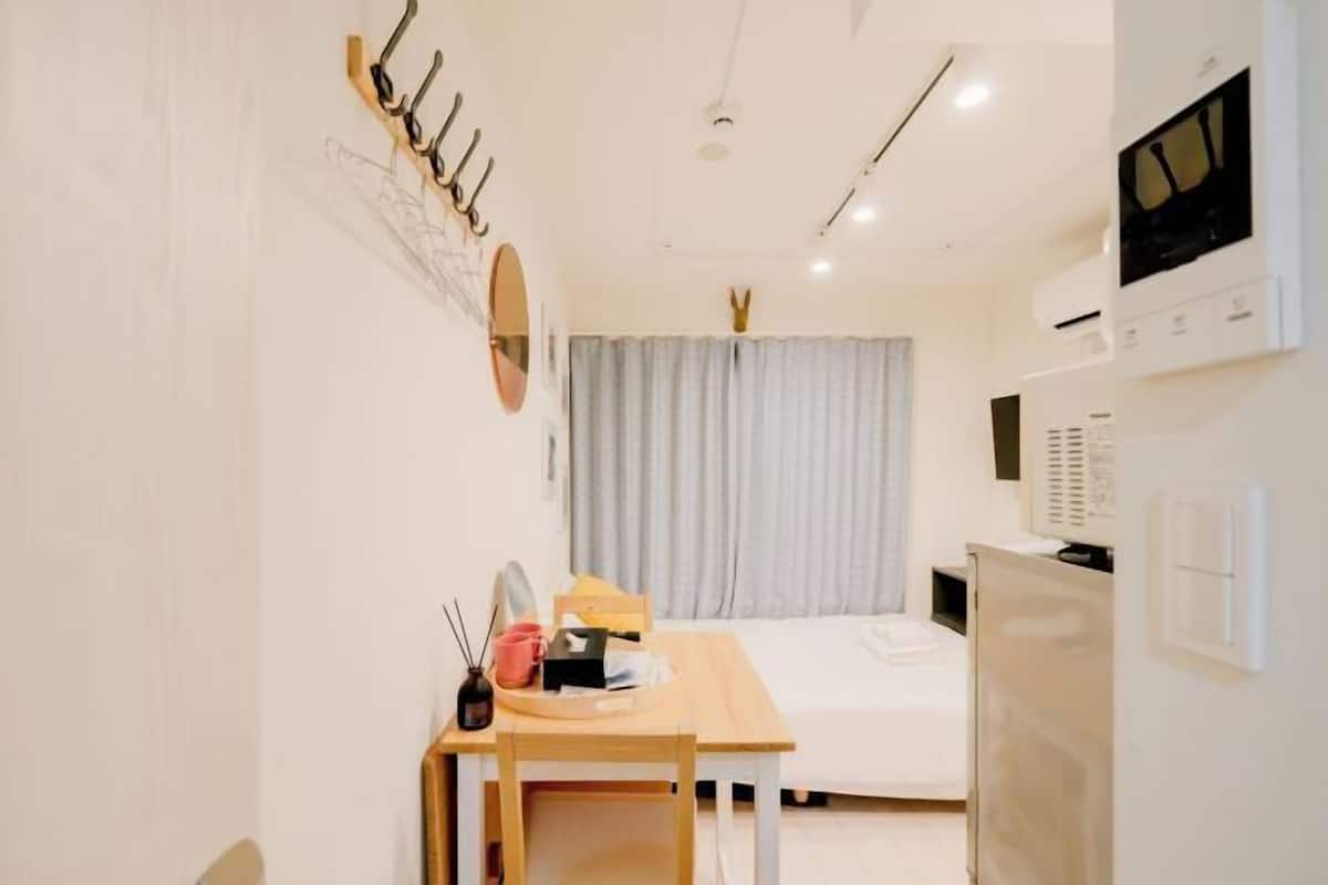 A compact hotel room with a bed, a desk with a chair, a mirror, a TV, and a coat rack.