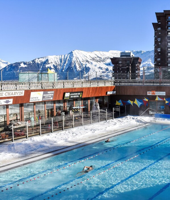 Una piscina con varias pistas, rodeada de edificios y montañas nevadas al fondo.