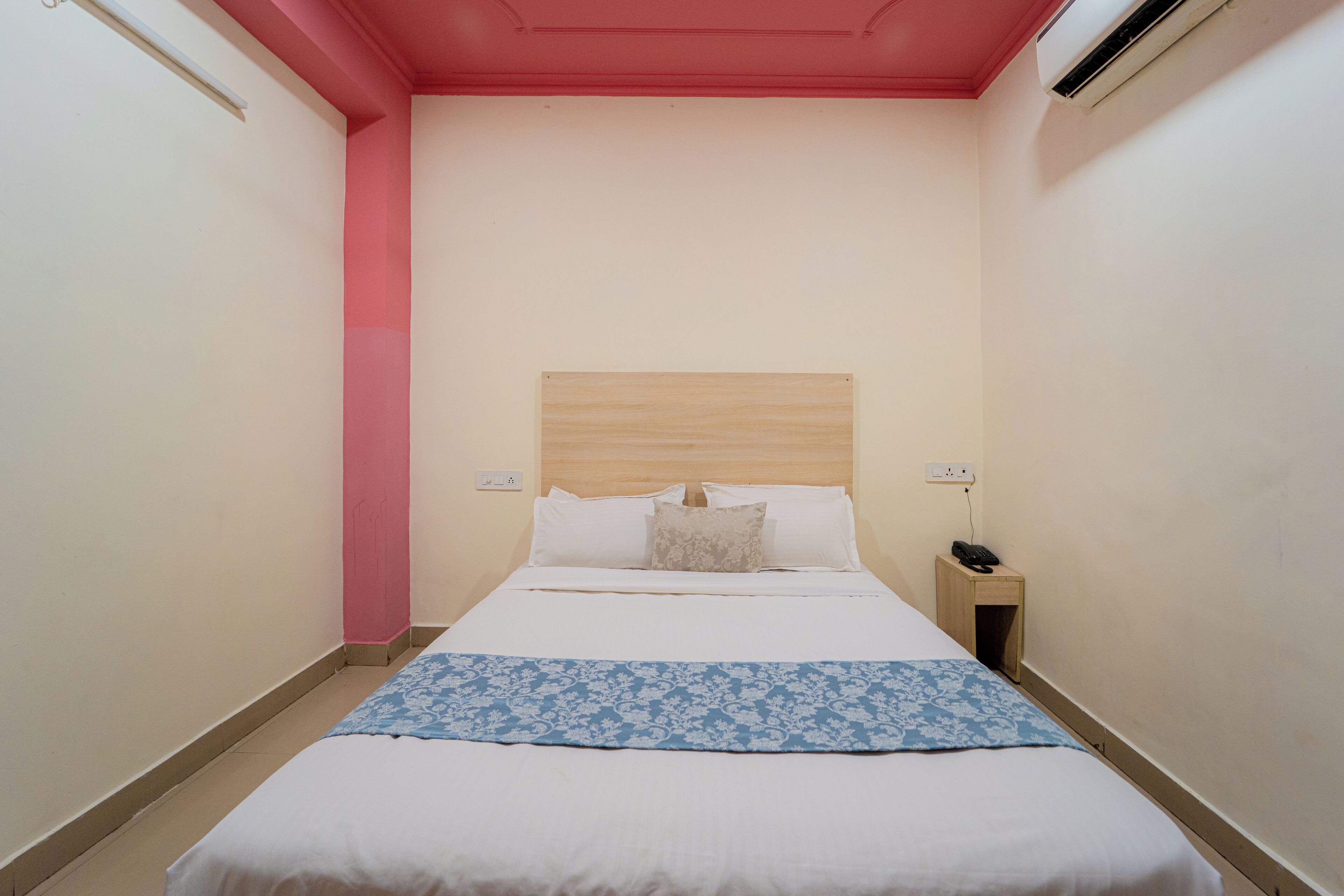 A narrow hotel room with a single bed, a bedside table, and a nightstand with a phone.