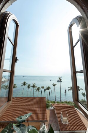 Vista desde una habitación con piso de baldosas y ventana con vistas a una playa con palmeras y barcos.