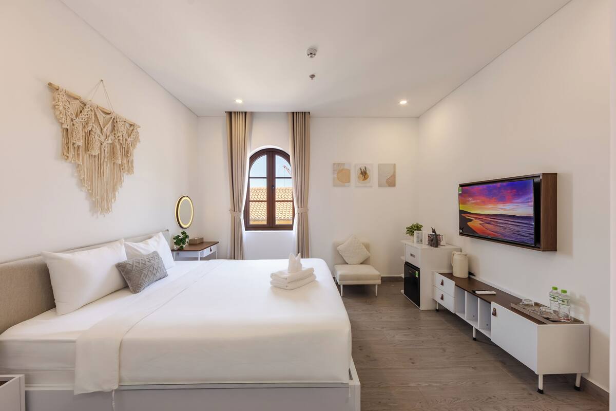 A modern hotel room with a large bed, a TV, a small seating area, and a view through a window.
