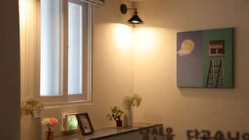 A room with a window, a wall-mounted light, a framed picture, and a small shelf with decorative items.
