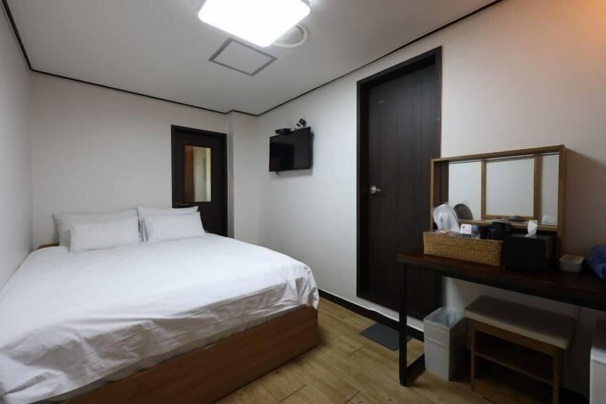 A bedroom with a bed, a desk with a mirror, a door, and a small trash bin.
