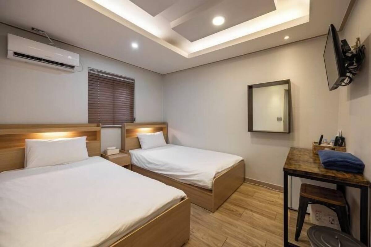 A hotel room with two beds, a desk, a chair, an air conditioning unit, a window with blinds, and a mirror on the wall.
