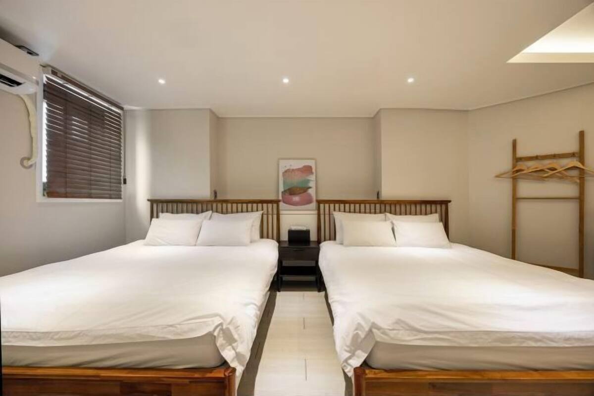 A hotel room with two single beds, a wooden headboard, a bedside table, a wall-mounted air conditioning unit, a window with blinds, and a hanging wooden rack.