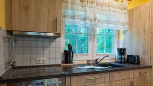 A kitchen with wooden cabinets, a stainless steel stove, a window with curtains, and a coffee maker. - Am Deich Haus 32 - Comfortable vacation home on the dike with terrace, garden and (Zingst)