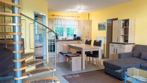 A modern kitchen with a central island, wooden cabinets, and a dining area with a table and chairs. There is a staircase with a metal railing leading to an upper level. - Am Deich Haus 32 - Comfortable vacation home on the dike with terrace, garden and (Zingst)