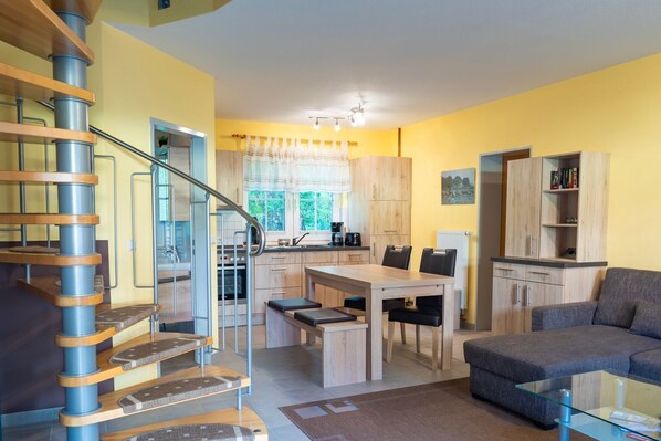 Une cuisine moderne dotée d’un îlot central, de meubles en bois et d’un coin repas avec une table et des chaises. Un escalier avec une rampe en métal mène à l’étage supérieur.