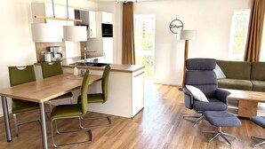 Una cocina moderna con armarios blancos, encimera de madera y un comedor con mesa de madera y sillas verdes. Hay un sofá gris y una lámpara de pie en la zona de estar.