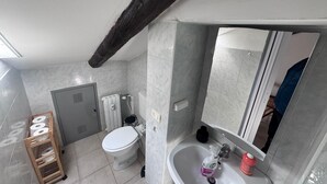 Ein Badezimmer mit Toilette, Waschbecken und Spiegel. Ein Holzregal mit Toilettenpapierrollen und einem Heizkörper.