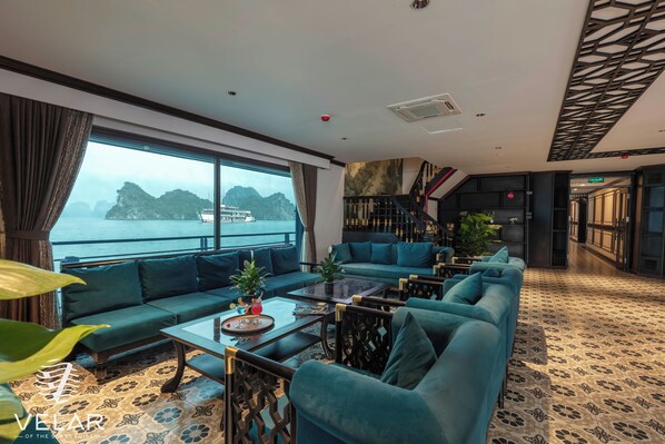 A modern lounge with a sofa, armchairs, and a coffee table. A large window offers a view of the sea and distant islands. - Velar Of The Sea Cruise (Tuan Chau)