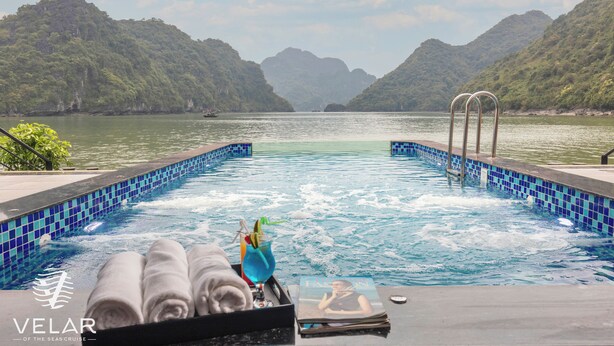 A pool with a view of a lake and mountains, towels, and a drink on the edge. - Velar Of The Sea Cruise (Tuan Chau)