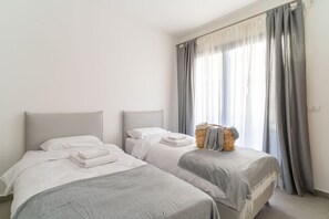 A room with two beds, a window with curtains, and a basket with towels. - Luxury 3 rooms Apt• Balcony• Parking | HaKerem A45 (680012)