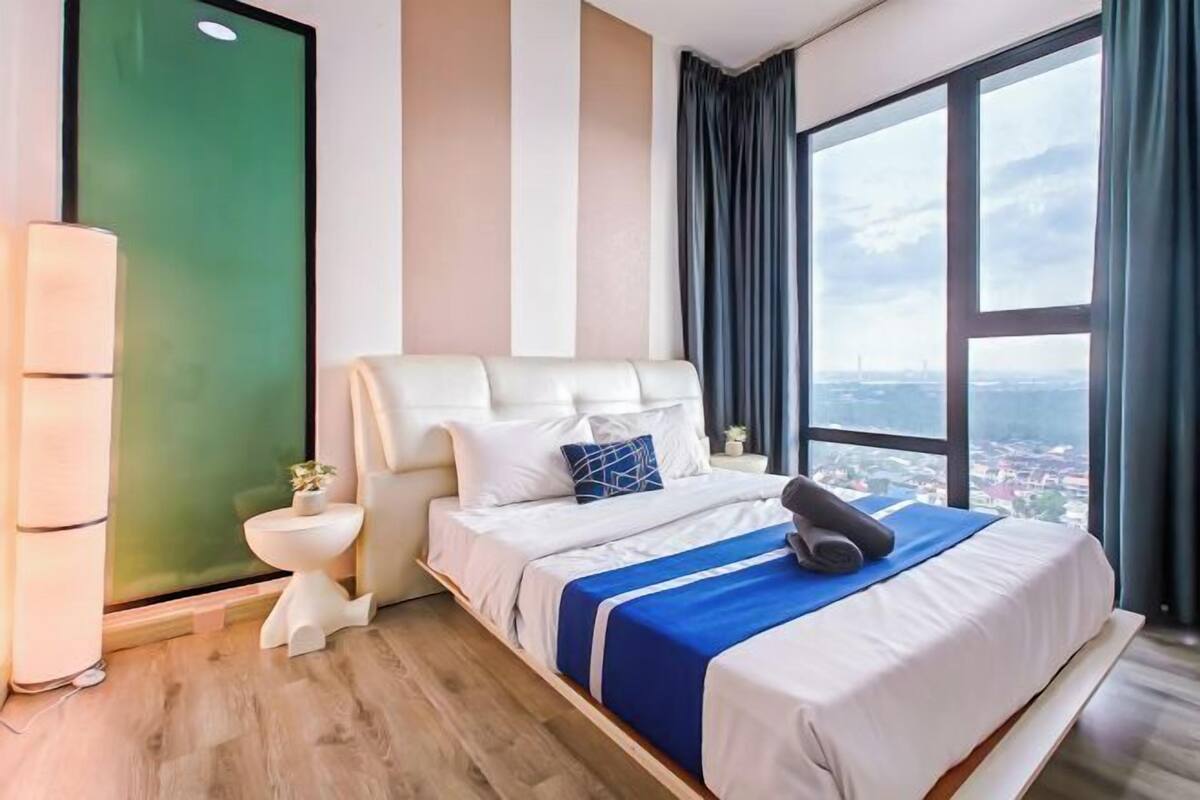 Een moderne hotelkamer met een groot bed, een klein nachtkastje, een groene deur en een stadszicht door grote ramen.