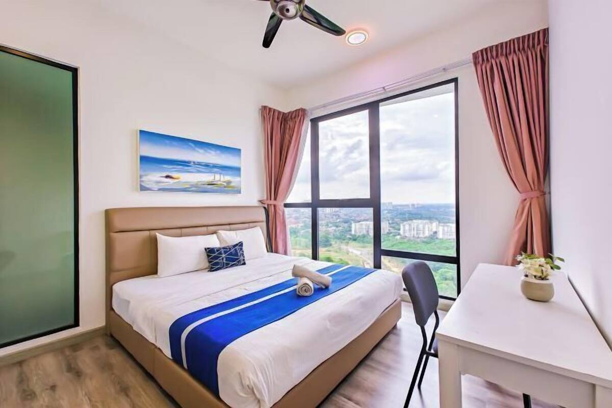 Hotelkamer met een bed, bureau, stoel, plafondventilator, raam met gordijnen en een schilderij aan de muur.