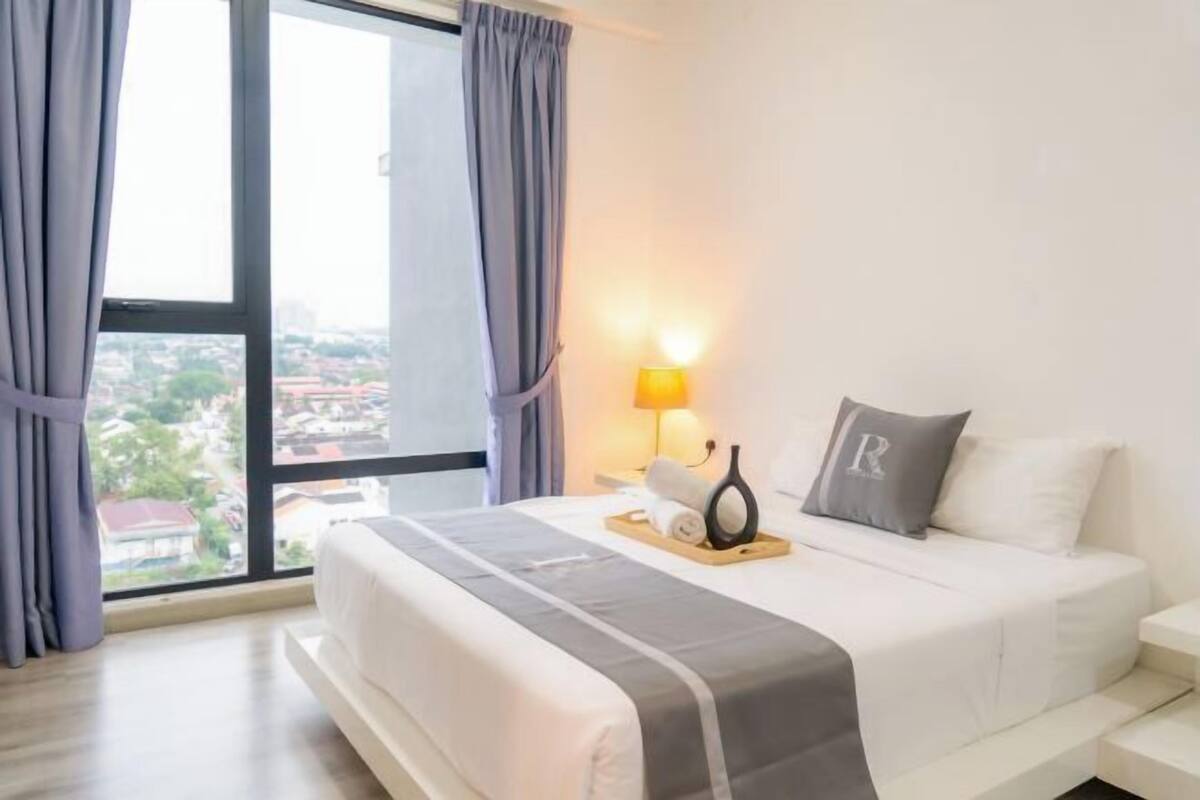 Hotelkamer met een groot raam, een bed met een grijze plaid, een nachtkastje met een lamp, en uitzicht op de stad.