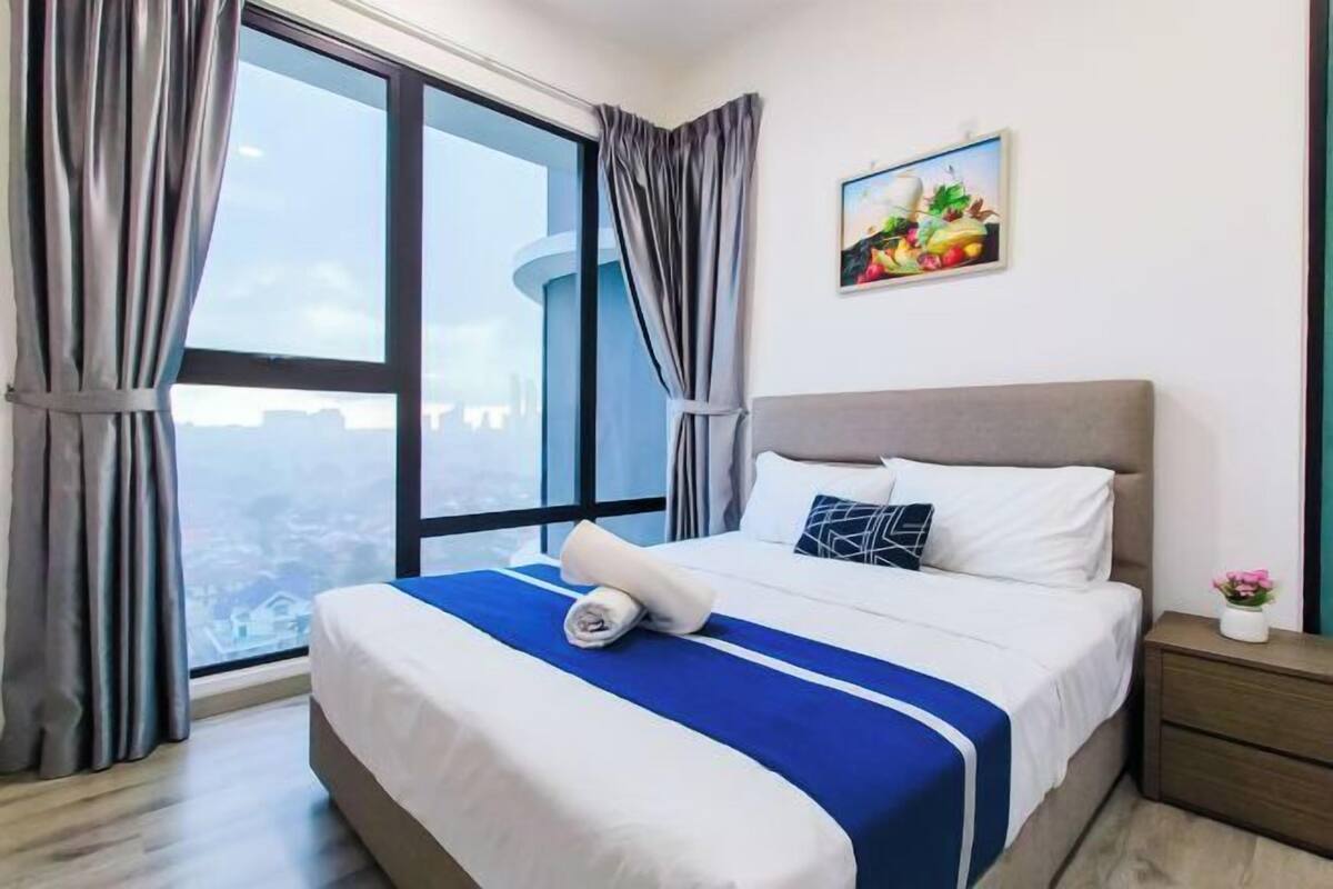 Een hotelkamer met een groot raam, een bed met een blauw-witte sprei, een nachtkastje met een vaas en een schilderij aan de muur.