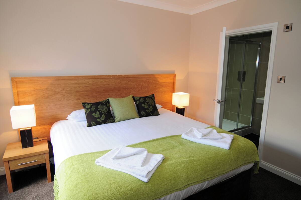 Habitación de hotel con una cama grande, dos lámparas de noche, una manta verde y una ducha con cerramiento de vidrio.